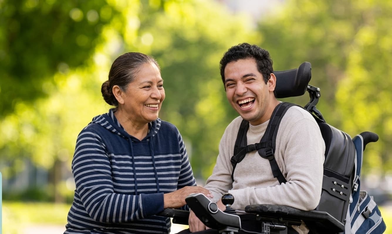 An older woman and a young man who uses a wheelchair laughing together outdoors